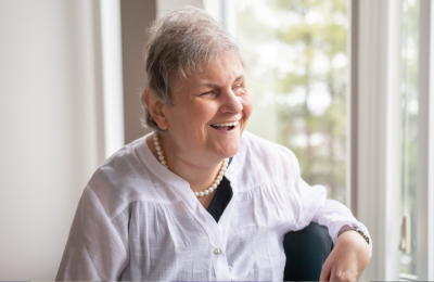A white older white woman adult wearing a blouse she is sitting in chair near window smiling.